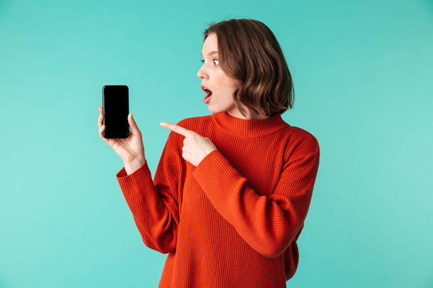Portrait of an excited young woman dressed in sweater pointing finger at blank screen mobile phone isolated over blue background - Fotó, kép