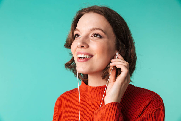 Portrait of a smiling young woman dressed in sweater listening to music in earphones isolated over blue background - Φωτογραφία, εικόνα