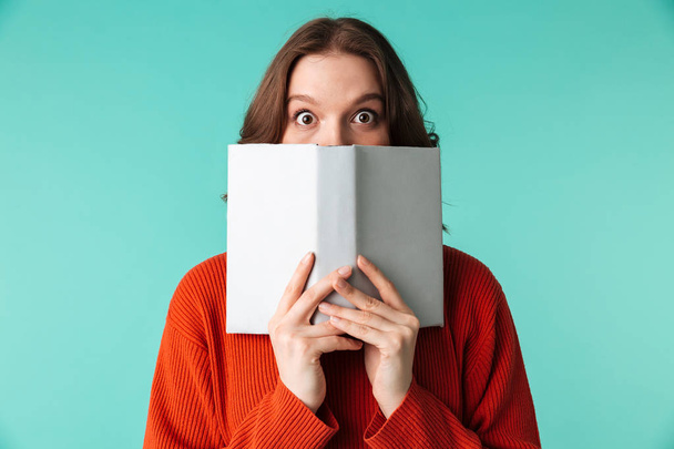 Portrait of a surprised young woman dressed in sweater holding book at her face isolated over blue background - Foto, immagini