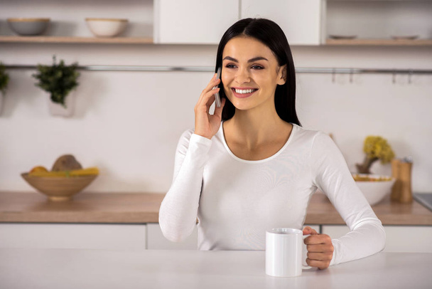 Full of gladness. Cheerful attractive young woman talking on smart phone while sitting in the kitchen - Foto, Imagem