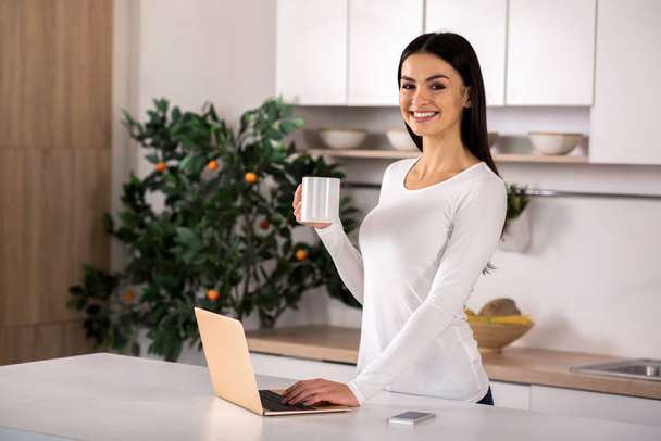 Pure joy. Waist up of nice woman standing in the kitchen while drinking tea and using laptop - Photo, image