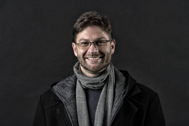 Fashion model with beard smile on dark background. Happy man in glasses on bearded face. Bearded man in scarf and coat on dark background. Autumn style and trend. Fashion accessory, vintage filter - Photo, image