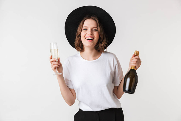 Portrait of a cheerful young woman dressed in black hat holding bottle of champagne and a glass isolated over white background - 写真・画像
