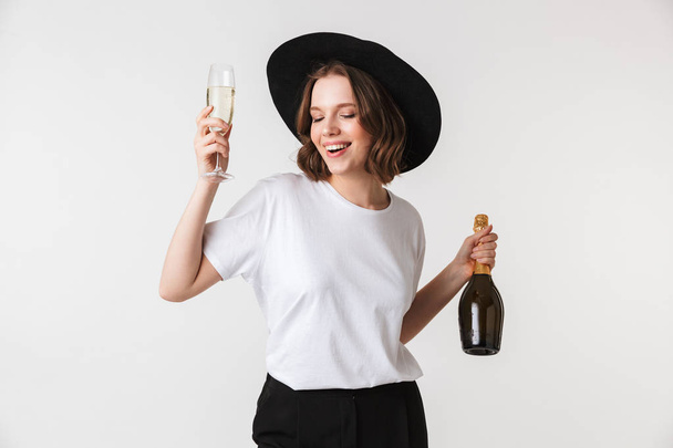 Portret van een gelukkige jonge vrouw gekleed in zwarte hoed holding fles champagne en een glas geïsoleerd op witte achtergrond - Foto, afbeelding