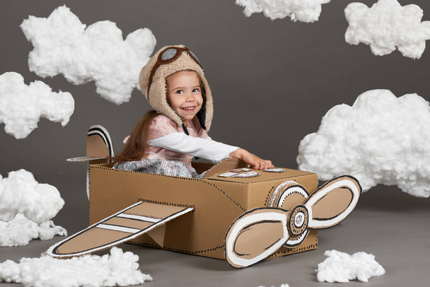 the child girl plays in an airplane made of cardboard box and dreams of becoming a pilot, clouds of cotton wool on a gray background - Zdjęcie, obraz