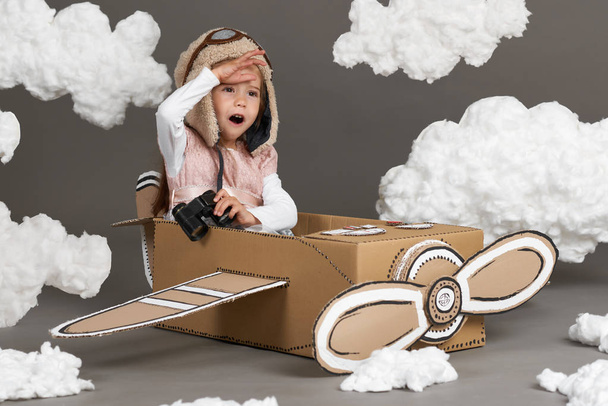 het kind meisje speelt in een vliegtuig gemaakt van kartonnen doos en droomt ervan om een piloot, wolken van katoen, wol op een grijze achtergrond - Foto, afbeelding