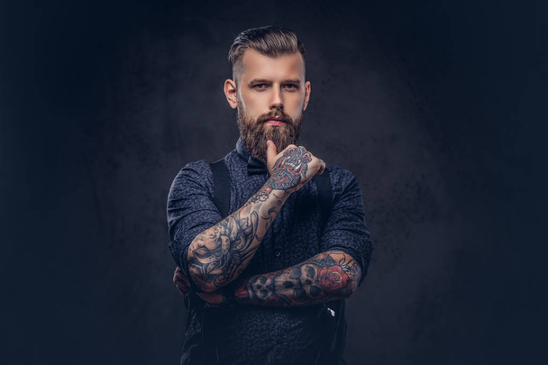 Pensativo guapo hipster anticuado en una camisa azul y tirantes, de pie con la mano en la barbilla en un estudio
. - Foto, imagen