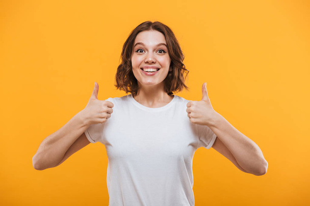 Image of excited young woman standing isolated over yellow background showing thumbs up gesture looking camera. - Fotó, kép