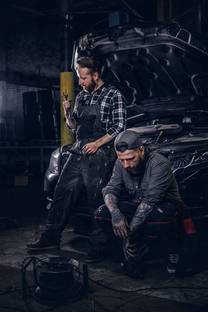 Dos mecánicos se sientan en un coche roto, descansan después de un duro día de trabajo en un garaje
. - Foto, imagen
