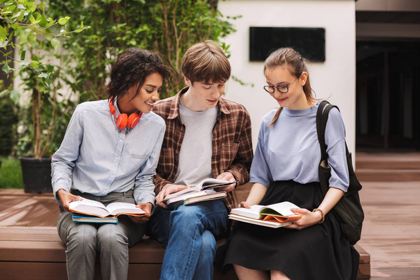 Group of smiling students sitting on bench and reading books in courtyard of university. Company of young students study together outdoors - Фото, изображение