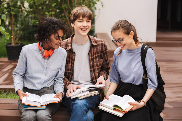 Группа счастливых студентов, сидящих на скамейке и читающих книги во дворе университета. Компания молодых улыбающихся студентов учится вместе на открытом воздухе
 - Фото, изображение