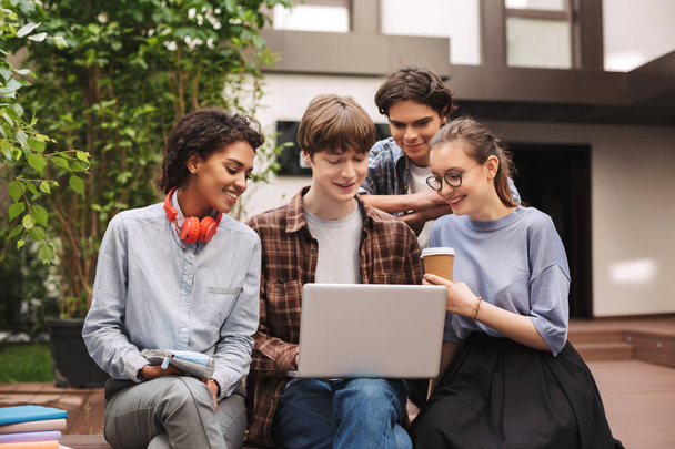 Groupe de jeunes étudiants souriants assis sur un banc et travaillant ensemble sur un ordinateur portable tout en passant du temps dans la cour de l'université. Compagnie d'étudiants étudiant en plein air
 - Photo, image