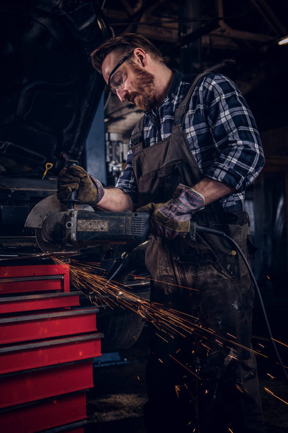 Brutal auto mechanic in a uniform and safety glasses working with an angle grinder while standing against a broken car in repair garage. - Photo, image