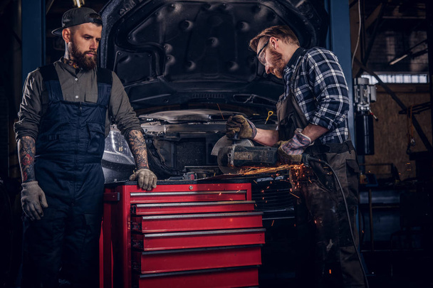 Zwei bärtige Automechaniker in Uniform und Schutzbrille arbeiten mit einem Winkelschleifer, während sie in der Werkstatt vor einem kaputten Auto stehen.  - Foto, Bild