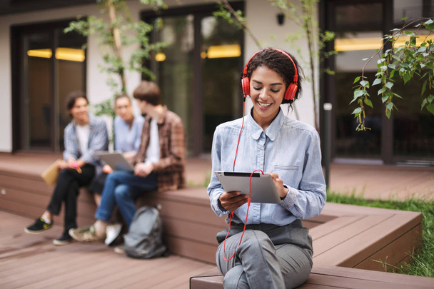 Ładna pani siedzi na ławce w czerwonej słuchawki i tablet w ręce na dziedzińcu Uniwersytetu z uczniami na tle - Zdjęcie, obraz
