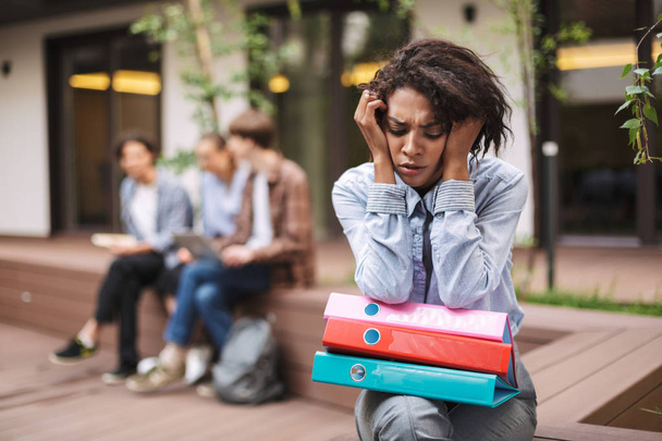 Портрет засмученої леді, що сидить на лавці з різнокольоровими папками на колінах і сумно закриває очі, проводячи час у дворі університету
 - Фото, зображення