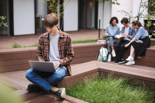 Young man sitting on bench and sadly working on laptop while spending time in courtyard of university with students on background - Zdjęcie, obraz
