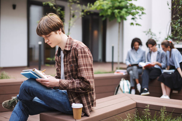 Νέοι στοχαστικός άνθρωπος κάθεται σε παγκάκι με καφέ και γράφοντας στο σημειωματάριο, ξοδεύοντας το χρόνο στο προαύλιο του Πανεπιστημίου με τους μαθητές σε φόντο - Φωτογραφία, εικόνα
