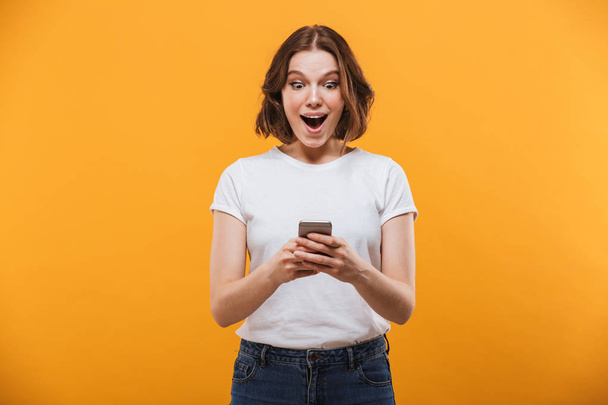Image of excited young woman standing isolated over yellow background chatting by mobile phone. Looking aside. - Photo, Image