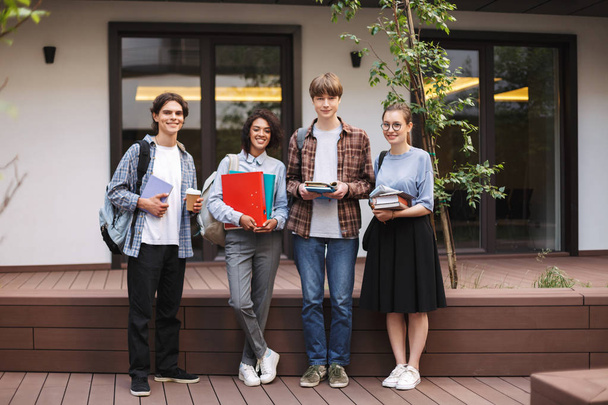 Молодые студенты стоят с книгами и папками в руках и радостно смотрят в камеру, проводя время вместе во дворе университета
  - Фото, изображение