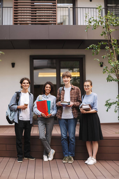 Jeunes étudiants souriants debout avec des livres et des dossiers dans les mains et regardant joyeusement à la caméra tout en passant du temps ensemble dans la cour de l'université
  - Photo, image