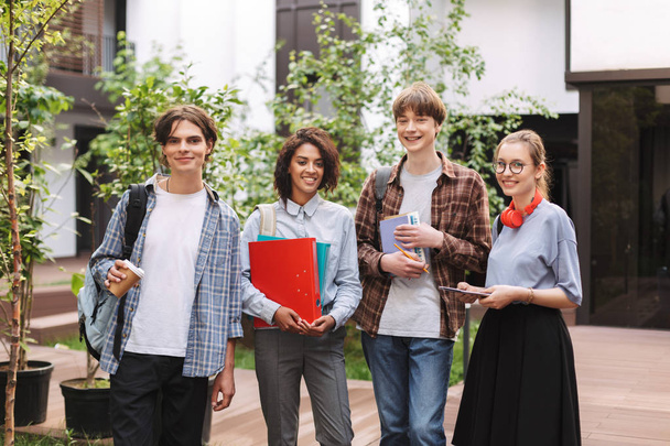 Grupo de jovens estudantes sorrindo em pé com livros e pastas nas mãos e olhando alegremente para a câmera enquanto passam o tempo juntos no pátio da universidade
  - Foto, Imagem