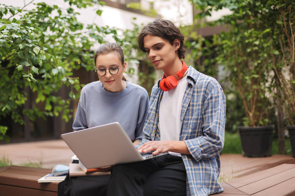 Δυο χαμογελώντας μαθητές κάθονται στον πάγκο και εργάζονται για το laptop στην αυλή του Πανεπιστημίου. Νεαρός άνδρας και κυρία σπουδάζοντας μαζί σε εξωτερικούς χώρους - Φωτογραφία, εικόνα