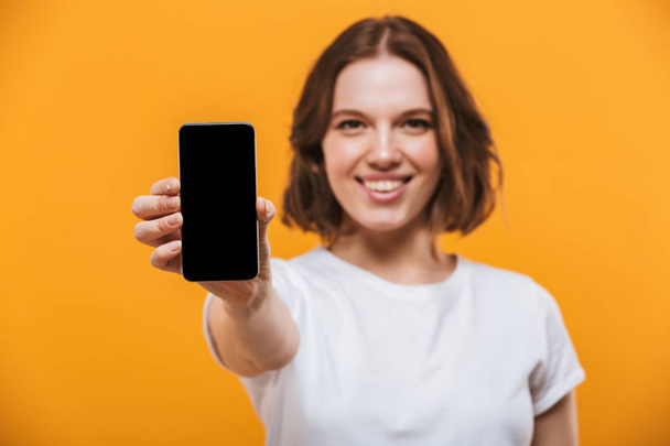 Photo of happy young woman standing isolated over yellow background showing display of by mobile phone. Looking camera. - Foto, immagini
