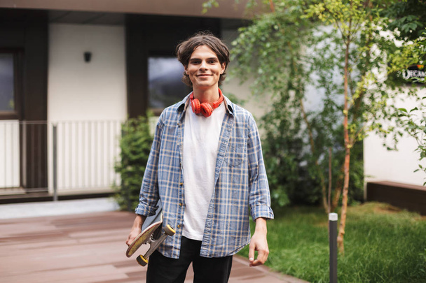 Niño sonriente con auriculares rojos de pie y sosteniendo el monopatín mientras mira alegremente en la cámara en el patio de la universidad
 - Foto, Imagen