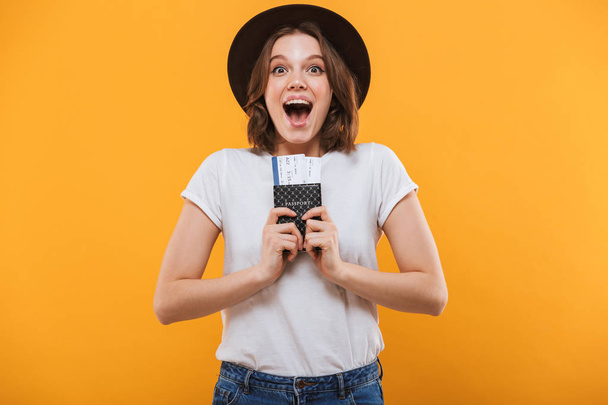 Image of excited emotional young woman tourist standing isolated over yellow background holding passport with tickets looking camera. - Photo, Image