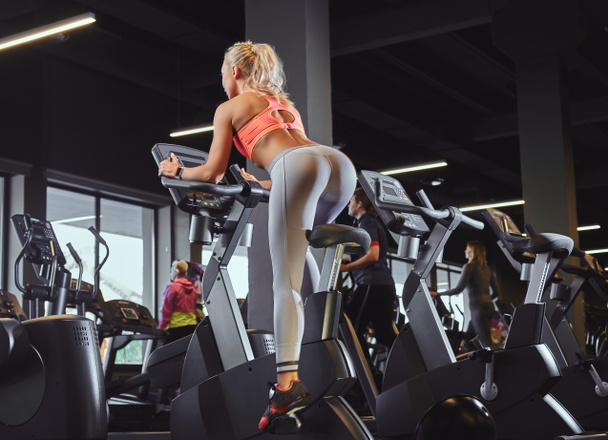 Jeune belle femme blonde en tenue de sport s'entraînant sur un vélo d'appartement dans un centre de fitness moderne
. - Photo, image