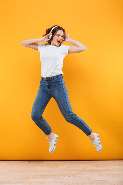 Image of emotional funny young woman jumping isolated over yellow background listening music. - 写真・画像