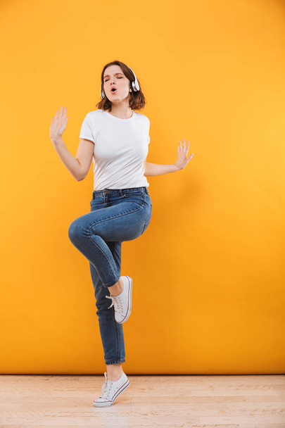 Full length portrait of a satisfied young woman in sweater listening to music with headphones and dancing over yellow background - Foto, immagini