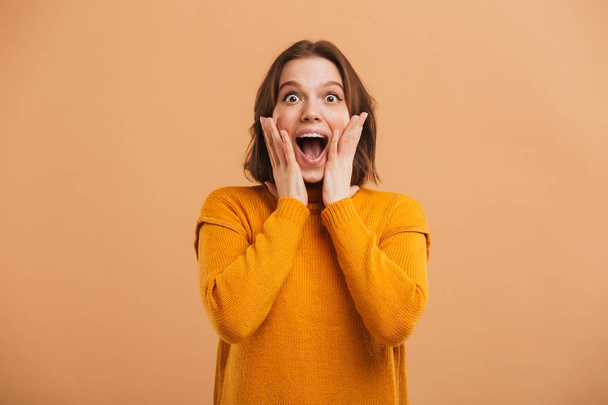 Portrait of an excited young woman in sweater celebrating success and screaming over beige background - Φωτογραφία, εικόνα