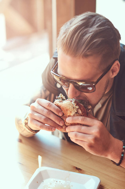 Portrait of a hungry hipster guy with a stylish haircut and beard sits at a table, decided to dine at a roadside cafe, eating a hamburger. - Foto, imagen
