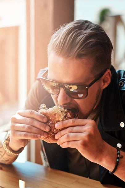 Nälkäisen hipsterin muotokuva, jolla on tyylikäs kampaus ja parta, istuu pöydässä, päätti syödä tien vieressä kahvilassa, syö hampurilaisen.
. - Valokuva, kuva