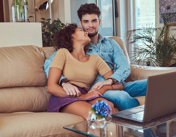 Feliz casal atraente durante um fim de semana, assistindo filme no laptop sentado no sofá em casa
. - Foto, Imagem