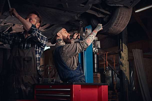 zwei bärtige Automechaniker in Uniform reparieren die Aufhängung der Autos, während sie unter dem Hubwagen in der Werkstatt stehen.  - Foto, Bild