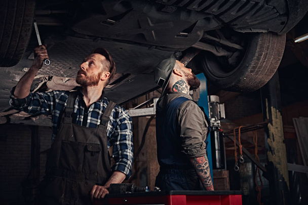Два бородатых автомеханика в униформе ремонтируют подвеску автомобилей, стоя под подъемником в ремонтном гараже
.  - Фото, изображение