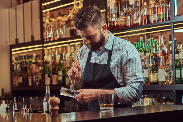 Стильный бородатый бармен в рубашке и фартуке раскалывает лёд, чтобы сделать коктейль на заднем плане барной стойки
. - Фото, изображение