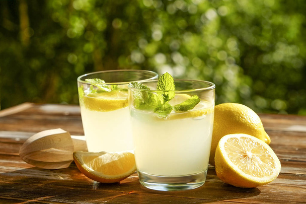 Two glasses full of ice cold refreshing lemonade beverage w/ mint leaves in sunlight on brown grunged wooden table, juicer, ripe organic lemon, country side foliage background. Copy space, close up. - Фото, зображення