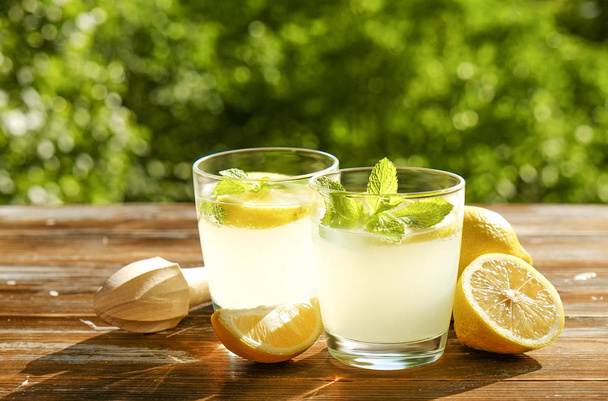 Two glasses full of ice cold refreshing lemonade beverage w/ mint leaves in sunlight on brown grunged wooden table, slices of ripe organic lemon, country side foliage background. Copy space, close up. - Foto, afbeelding