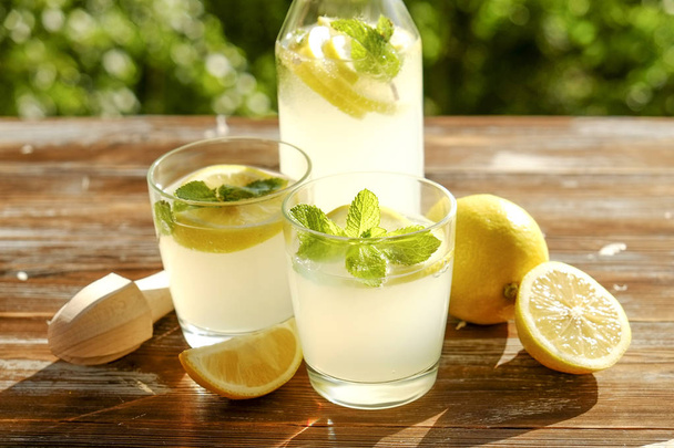 Lemonade pitcher, juicer, two glasses full of ice cold refreshing citrus beverage with lemon slices & mint leaves brown grunged wooden table, country side foliage background. Close up, copy space. - Фото, зображення