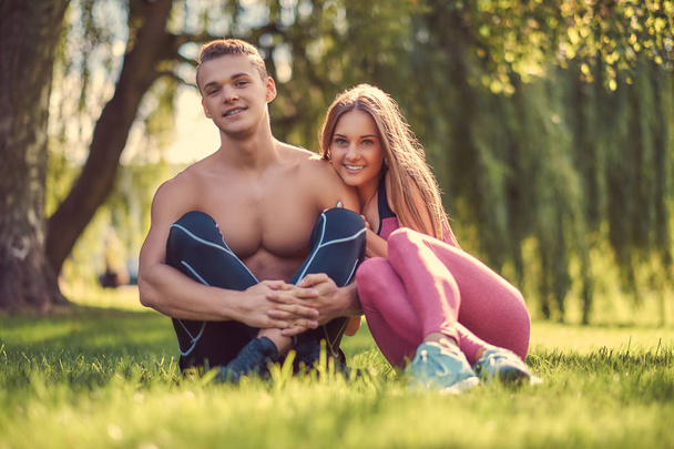 健康的なライフ スタイルのコンセプト。緑の芝生に座って抱きしめる幸せな若いフィットネス カップル. - 写真・画像