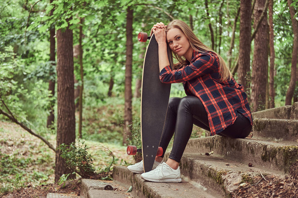 Young blonde hipster girl resting in a park, after riding on the skateboard. - Photo, Image
