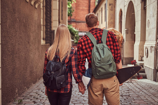 Achteraanzicht van een paar jonge hipster, knappe schaatser en zijn vriendin, hand in hand, lopen rond een oude smalle straatjes van Europa. - Foto, afbeelding