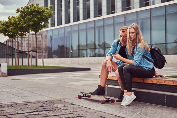 Giovane coppia attraente felice in abiti casual seduto su una panchina contro un grattacielo, guardando qualcosa su uno smartphone
. - Foto, immagini