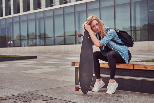 Hipster νεαρή ξανθιά κοπέλα σε casual clothessitting σ ' ένα παγκάκι εναντίον ενός ουρανοξύστη, αναπαύεται μετά την ιππασία για skateboard. - Φωτογραφία, εικόνα
