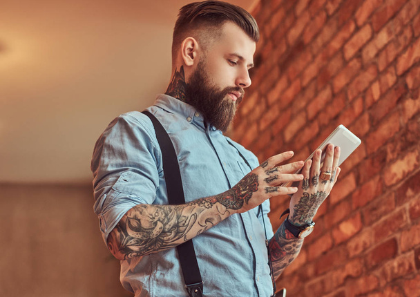 Ντεμοντέ τατουάζ hipster φορώντας ένα πουκάμισο και ζαρτιέρες, σε ένα γυαλιά ηλίου, χρησιμοποιώντας ένα tablet ενώ στέκεται κοντά σε ένα γραφείο σε γραφείο με εσωτερικό πατάρι. - Φωτογραφία, εικόνα