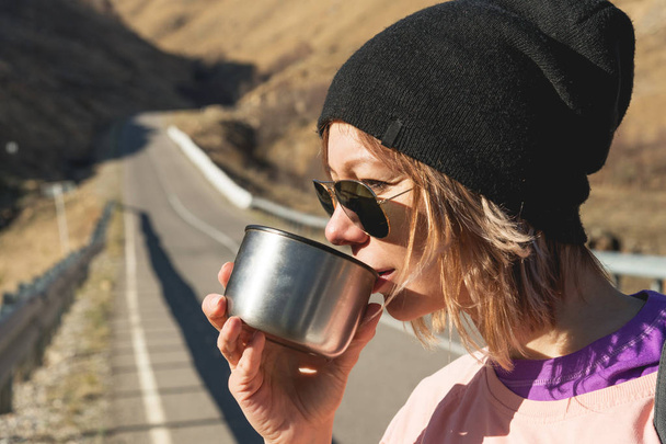 A hipster girl wearing sunglasses and a black hat is drinking tea or coffee from a mug from a thermos in the wild on a journey - Fotografie, Obrázek